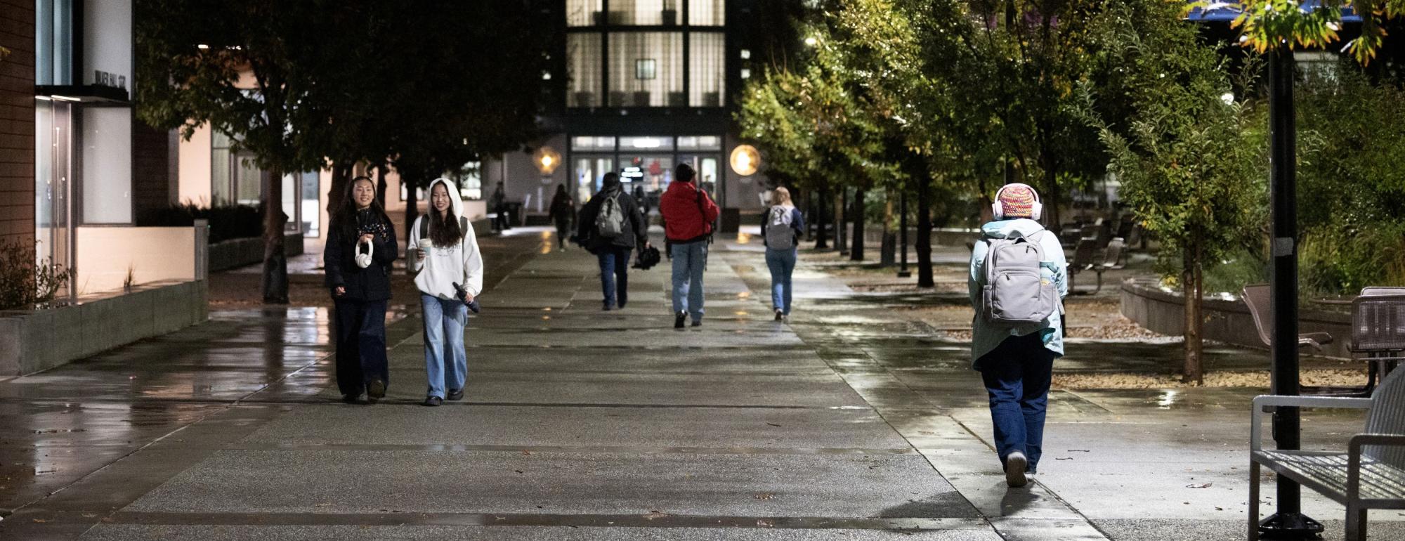 Students walking along a wet path lined with trees at night, the glow of Shields Library in the background.