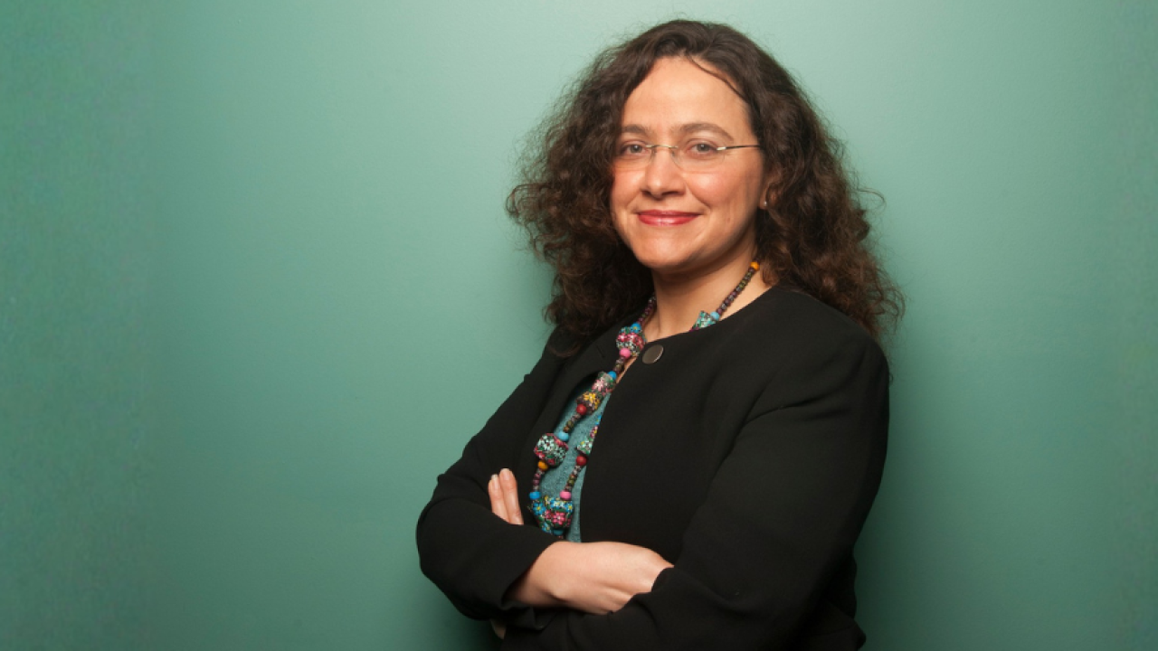 Smiling woman with curly hair and glasses in black blazer, arms crossed.