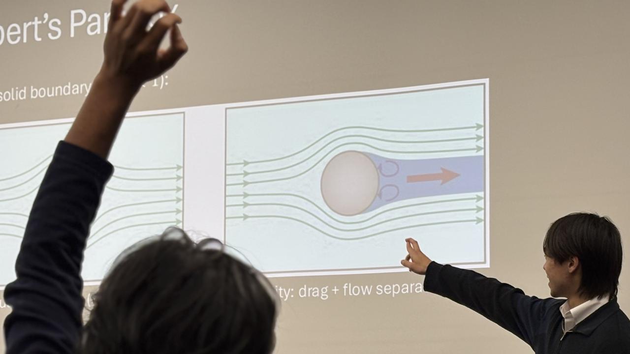 A person gestures towards a presentation on fluid dynamics, explaining concepts with diagrams.