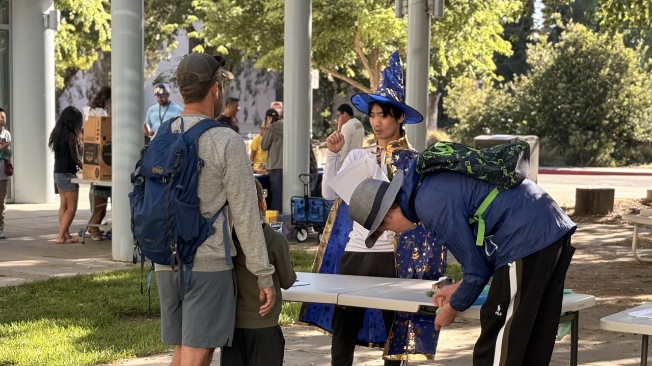 A student in a blue wizards hat explains mathemagical tricks to several Picnic Day goers.