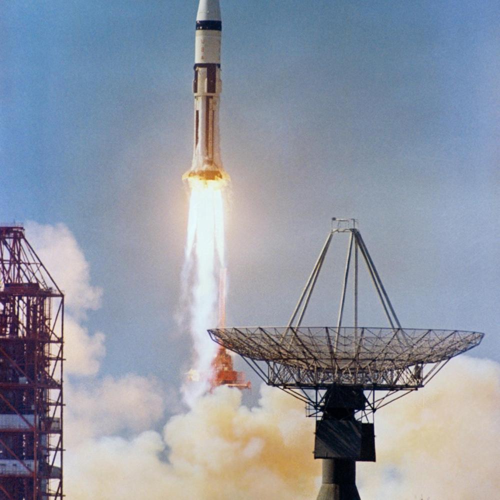 The Apollo 7 rocket launches from a cloud of smoke, with radar dishes in foreground.