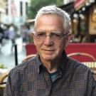 Craig Tracy, an older man with glasses sitting outdoors at a café, wearing a dark grey collared shirt, smiling softly.