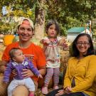 A casual family photo of Swati and Axel smiling with their two kids at a pumpkin patch. One child reaches out, while the baby sits on their father's lap looking confused.