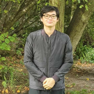 Nick Nho stands with hand clasped in front of him, a green lightly forested area stretches behind, with leaves on the ground.